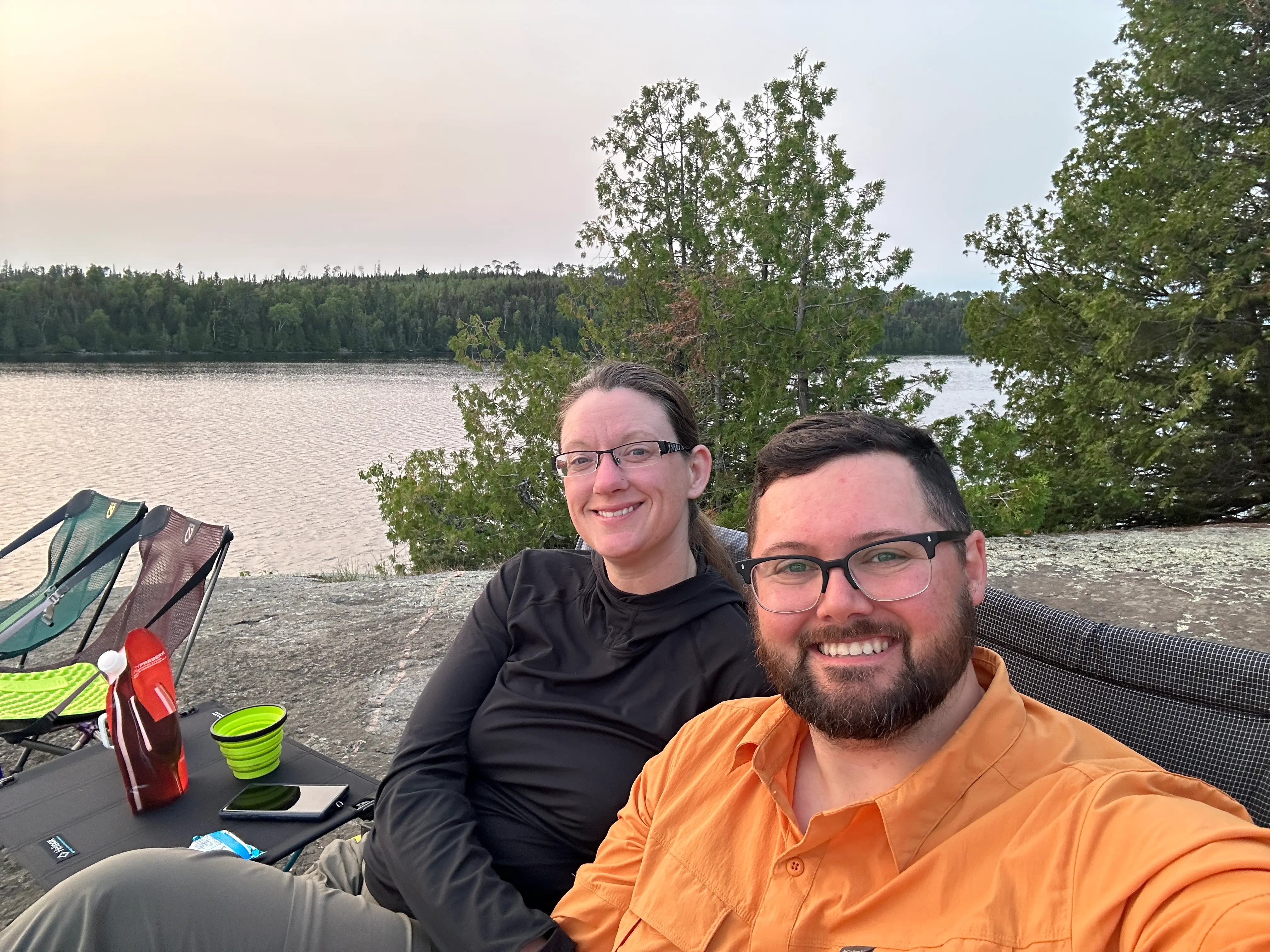 Paul & Annica sitting in camp chairs on a rock bluff overlooking a lake peppered with small evergreen trees